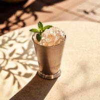 A Mint Julep cocktail captured from a slightly elevated angle, positioned on a round table in a Mediterrean patio. The Mint Julep cocktail is garnished with a fresh mint sprig. The scene creates the inviting atmosphere of a summer afternoon, bathed in warm sunlight that casts shadows of tree branches on the table and floor.